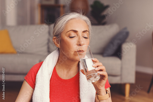 Cuadro en lienzo Focused Senior Woman Hydrating By Drinking Water After Workout At Home, Sitting In Living Room With Towel Around Neck, Promoting Wellness And Fitness Lifestyle