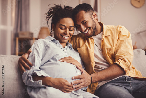 Obraz na płótnie Joyful African American Couple Smiling At Each Other While Enjoying Time Together In Cozy Living Room, Embracing Pregnant Belly In Maternity Celebration