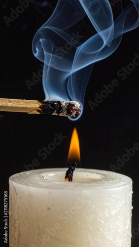 Close up of a burning match lighting a white candle flame with ethereal smoke trails against a dark background