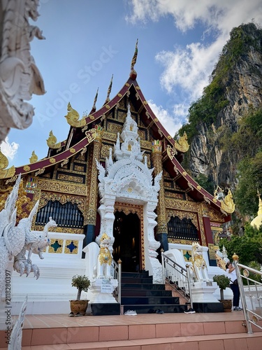 Detailed facade and entrance of a traditional Lanna-style temple building with intricate white wood carvings and a dramatic mountain cliff background at Wat Tham Chetawan in Nan province, Thailand.
