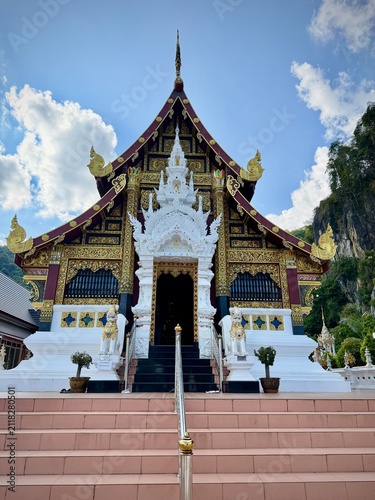 Detailed facade and entrance of a traditional Lanna-style temple building with intricate white wood carvings and a dramatic mountain cliff background at Wat Tham Chetawan in Nan province, Thailand.
