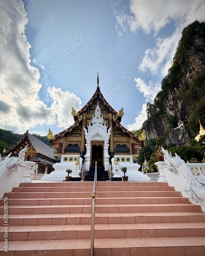 Detailed facade and entrance of a traditional Lanna-style temple building with intricate white wood carvings and a dramatic mountain cliff background at Wat Tham Chetawan in Nan province, Thailand.