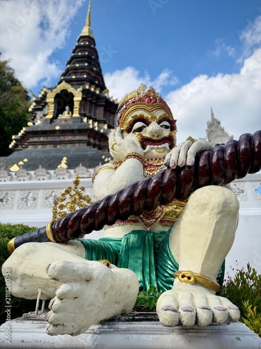 Close-up of a colorful giant demon guardian sculpture sitting with a club in front of an ornate tiered temple pagoda at Wat Tham Chetawan in Nan province, Thailand.