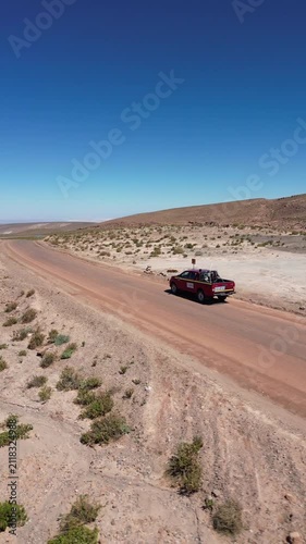 Orbit shot around red truck parked on winding desert road surrounded by dry hills and scattered scrubland under clear blue sky