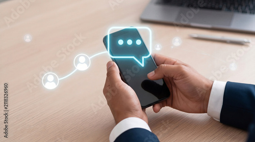 Businessman holding a smartphone with glowing chat and network icons on a wooden desk