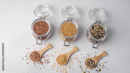 Three glass jars filled with different types of seeds and wooden spoons on a white background