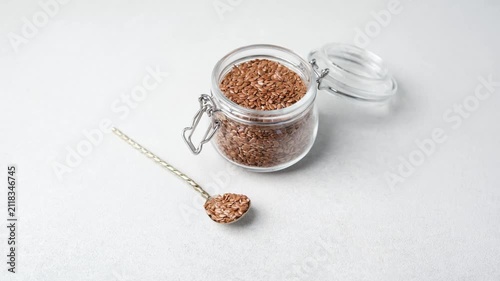 Small glass jar filled with flax seeds beside a spoon of seeds on a light gray background