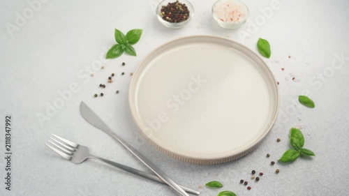 A clean plate with fork and knife surrounded by basil leaves and scattered peppercorns and salt