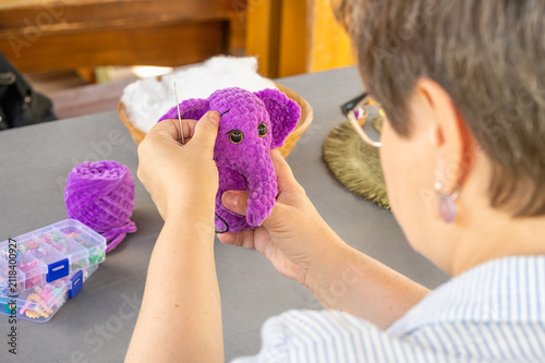 Woman stitching detail on purple plush crochet amigurumi elephant toy at work table. Cute stuffed animal crafting process. Unique custom handmade gift creation