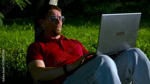 Busy man working on a laptop while sitting on the grass in a city park sitting under tree.