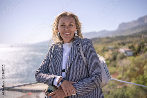 Принти на полотні Woman blazer traveler smiles by the sea railing with mountain views and bright s
