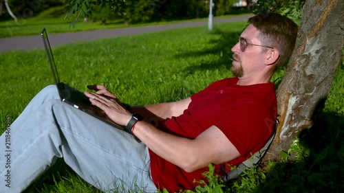 Busy man working on a laptop while sitting on the grass in a city park sitting under tree.