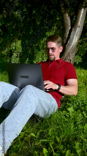 Busy man working on a laptop while sitting on the grass in a city park sitting under tree.