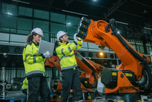 Industrial engineers team inspecting orange robotic arm automated machinery stock in warehouse factory floor. Industrial maintenance service, equipment quality inspection, smart manufacturing.