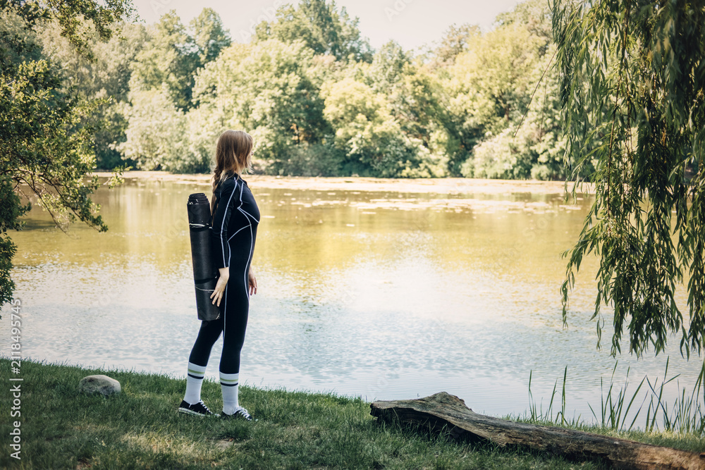 Fototapeta premium Woman with yoga mat standing by lake, contemplative nature walk, solo fitness journey, scenic workout motivation, peace and solitude