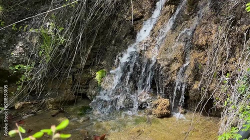 Close up of a natural forest waterfall cascading over textured rocks with clear flowing water, moss and woodland vegetation in bright summer sunlight. High quality 4k footage