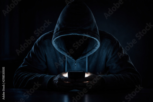 Mysterious hooded figure illuminated by a smartphone screen in darkness, representing cybercrime, online anonymity, hacking, and digital security threats.