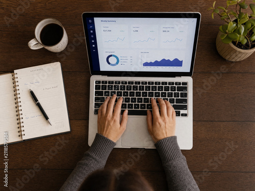 Person analysing business charts on a laptop at a wooden desk with coffee and a notepad