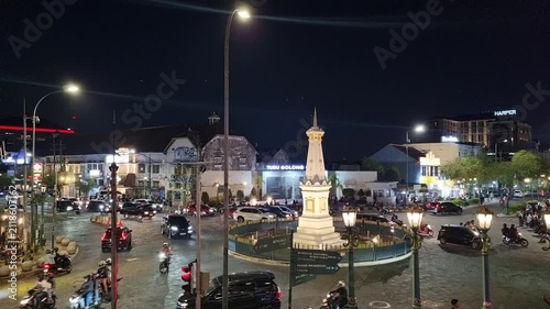 Real-time footage of the illuminated Tugu Yogyakarta landmark in Central Java, Indonesia, surrounded by lively evening street traffic, motorcycles, and cars navigating under bright city lights.