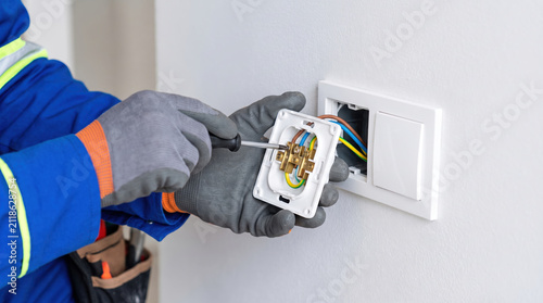 An electrician uses a screwdriver to work on a wall outlet in a home. The task involves handling wires and tools for repair or installation during daytime