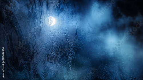 A bright moonlight glows through a foggy glass, creating a mystical scene at night. The glass is covered in water droplets and soft blue light surrounds the moon