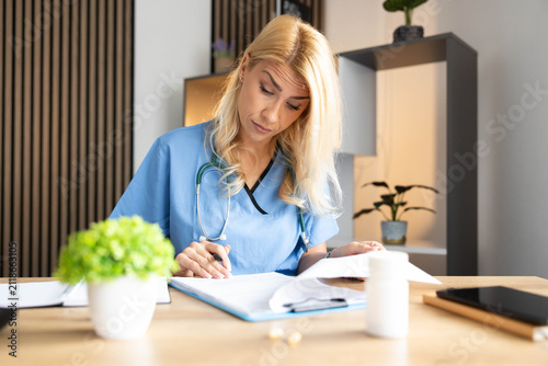 Female healthcare worker in scrubs and stethoscope concentrating on paperwork, managing essential administrative tasks at her office desk