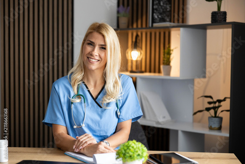 Confident female doctor wearing blue scrubs and stethoscope, sitting at a desk in a modern medical clinic, smiling