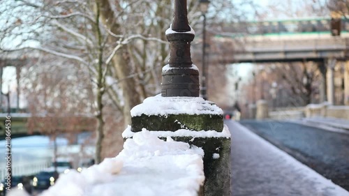 Winter scene in Paris with a close-up of a traditional lamp post covered in snow along a cobblestone street. A car with headlights on is driving in the background