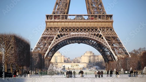Wide view of the Eiffel Tower with its base surrounded by snow-covered trees and park grounds in Paris. Captured on a bright winter morning, with people walking in the distance and long shadows.