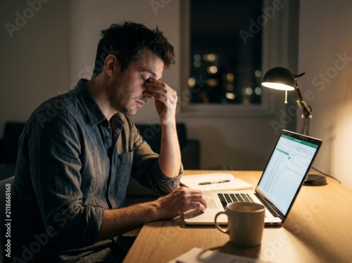 Exhausted man rubbing eyes while working late at night on laptop.