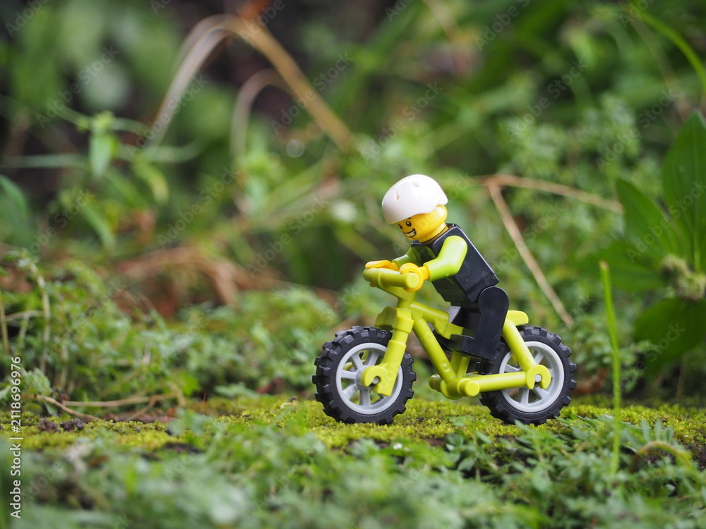 Fototapeta premium Indonesia - May 14, 2022 : Mini toy of action figure at grassland with blurred background. Miniature photography concept. Bicycle at outdoor with green grass conceptual design.