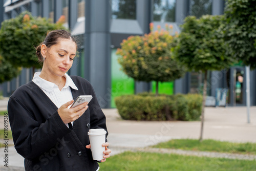 A focused businesswoman looking at her smartphone and holding a takeaway coffee cup while standing outdoors near an office complex, illustrating a busy urban lifestyle concept.