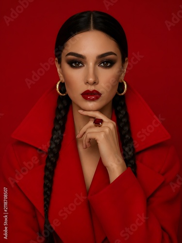 Centered fashion portrait of a woman wearing a sculptural red coat with wide collar, twin braids, gold hoop earrings and dramatic red lipstick.