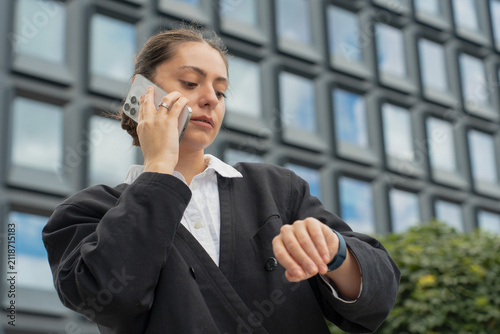 A businesswoman talking on her smartphone and checking her smartwatch outdoors against a modern office building, representing a time management and mobile communication concept.