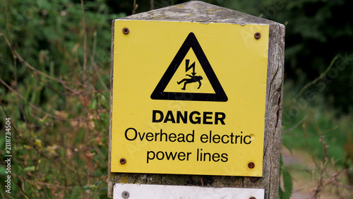 Danger overhead electric power lines warning sign, yellow high voltage risk notice on wooden post outdoors