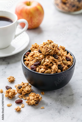 Healthy oat granola with raisins and pumpkin seeds in a black bowl with black coffee cup and fresh apple on marble table