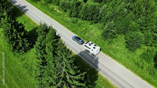 Car towing a travel trailer along a tree-lined roadside under bright summer sunlight