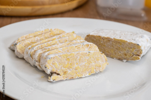 Sliced raw organic tempeh block on a clean white plate. Close up view showing the rich texture of fermented soybeans bound together by white mycelium. A healthy, plant-based vegan protein
