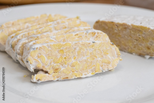 Sliced raw organic tempeh block on a clean white plate. Close up view showing the rich texture of fermented soybeans bound together by white mycelium. A healthy, plant-based vegan protein