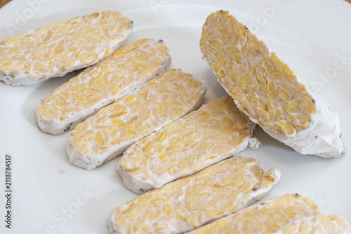 Sliced raw organic tempeh block on a clean white plate. Close up view showing the rich texture of fermented soybeans bound together by white mycelium. A healthy, plant-based vegan protein