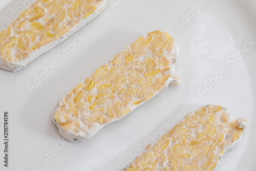 Sliced raw organic tempeh block on a clean white plate. Close up view showing the rich texture of fermented soybeans bound together by white mycelium. A healthy, plant-based vegan protein