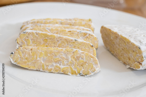 Sliced raw organic tempeh block on a clean white plate. Close up view showing the rich texture of fermented soybeans bound together by white mycelium. A healthy, plant-based vegan protein