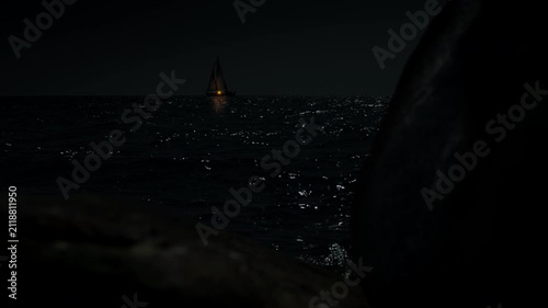 Sailboat sailing at night with a faint yellowish light surrounded by darkness with moonlight reflecting in the soft waves	