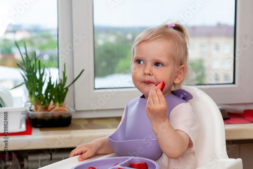 A young child sits at a table and enjoys fresh strawberries. Family members are present and smiling. The sun is shining through the window, creating a warm atmosphere.