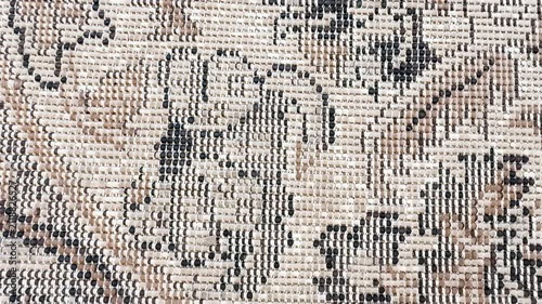 Detailed close-up of an aged woven carpet featuring a traditional geometric pattern in beige, cream, black, and brown tones. The camera slowly moves across the textured surface