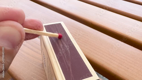Close-up of a hand striking a wooden match against the side of a matchbox on a wooden surface. The match ignites with a bright flame, burns for a moment, and then goes out, leaving a charred black tip