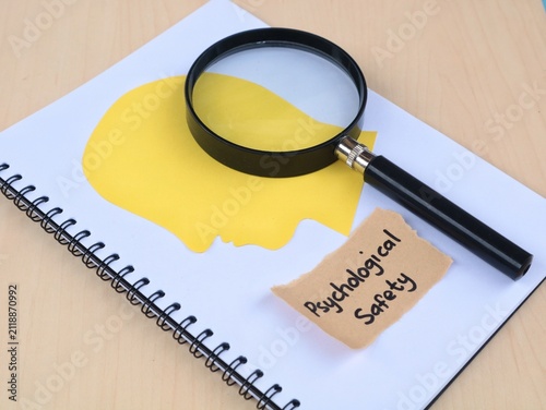 Psychological safety concept handwritten with human head paper silhouette and magnifying glass on book. Perfect for corporate culture, mental health at work, and human resource management concepts