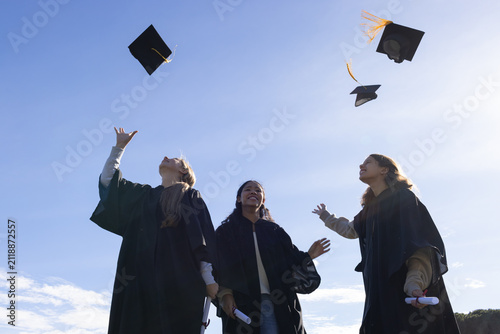 Three women graduates in gowns tossing tasseled caps on campus under blue sky, holding diplomas