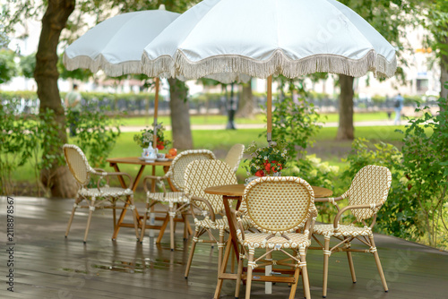 Outdoor cafe terrace with wicker chairs and wooden tables under vintage white umbrellas. Cozy summer restaurant veranda with elegant patio furniture setup in a park area.