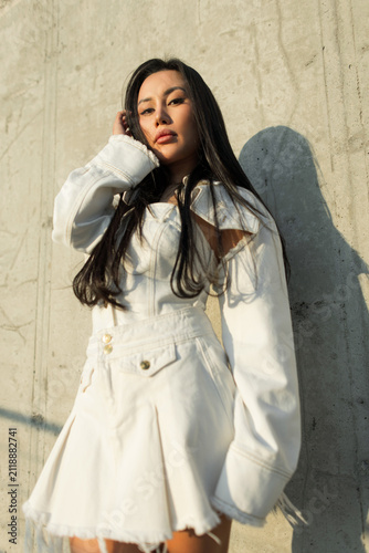 Obraz na płótnie Medium shot of a stunning young Asian woman in a trendy white denim outfit posing against a textured concrete wall in bright sunlight